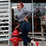 Woman in a checkered shirt and jeans poses next to a red fire hydrant, wearing red heels and a red bandana.