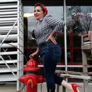Woman in a checkered shirt and jeans poses next to a red fire hydrant, wearing red heels and a red bandana.