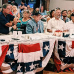 A group of people sit at a table decorated with patriotic bunting, attending an indoor event or celebration.