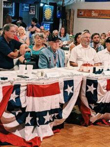 A group of people sit at a table decorated with patriotic bunting, attending an indoor event or celebration.